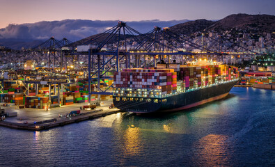 A illuminated container super cargo ship is being unloaded and loaded by cranes and trucks in a commercial terminal during evening