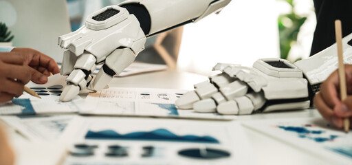 A close-up view of a robotic arm working alongside human hands in a collaborative business meeting, highlighting the integration of technology and strategy in modern workplaces.Noogenesis