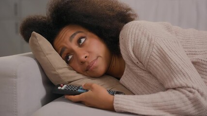African American tired woman feeling bad negative mood girl lying on sofa living room home holding remote controler switch channels watching television TV at home frustrated emotion exhausted female - Powered by Adobe