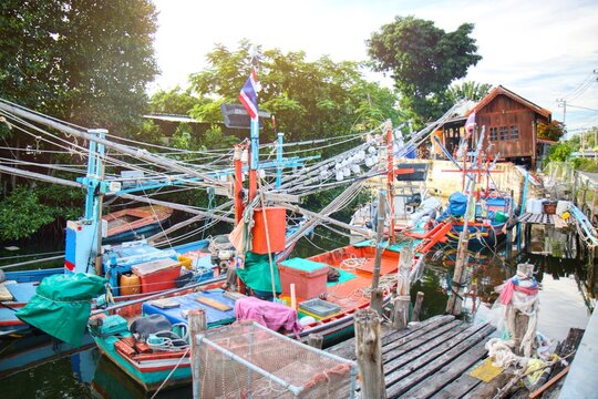 Traditional Thai fishing boats moored at a local pier in a coastal village. Authentic fisherman lifestyle featuring colorful wooden boats, fishing nets, and equipment under bright daylight. Scenic nau