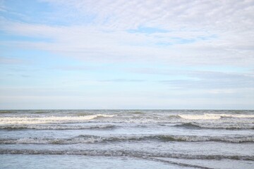 A serene and minimalistic coastal view of gentle ocean waves rolling onto the shore beneath a soft, pale blue sky with wispy white clouds.