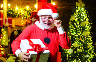 Smiling Santa Claus in glasses with Christmas gift box. Bearded man in Santa Claus wear with Christmas present. Festive New Year decorations. Christmas and New Year sales. Gift shop. Delivery service.