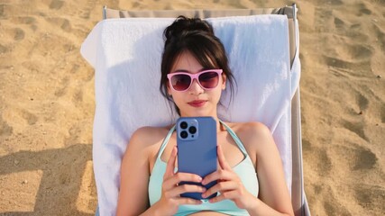 Top view of a beautiful young asian woman in a bikini and sunglasses relaxing on a beach chair, smiling and using her smartphone for social media while enjoying her summer vacation