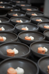 Numerous dark ceramic bowls are meticulously arranged, each containing an orange food roll and a dollop of white, herb-infused cream.
