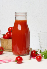 Glass bottle with tomato juice. Nearby are fresh red tomatoes and parsley leaves on a light background.