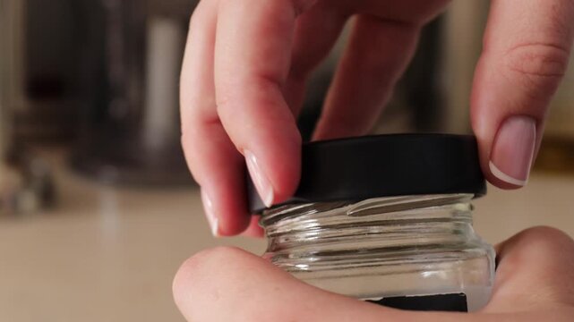 Hand gripping and rotating a silver metal lid on a transparent glass jar, performing a twisting motion to open or seal it in a home setting