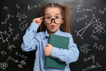Cute little child in glasses near black chalkboard with formulas and drawings