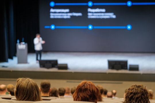 Conference stage audience listening to speaker with blue line graphics on large screen