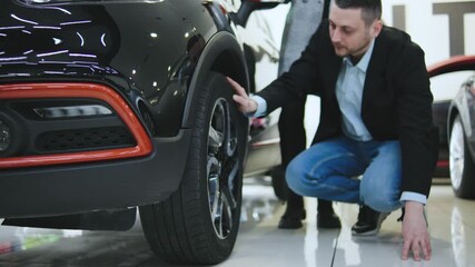 A client is kneeling beside a car in an automobile dealership, inspecting the wheels as a saleswoman offers guidance in a spacious sales salon.