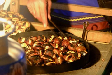 A view of chestnut kebab, a traditional Turkish street food, being cooked. A picnic lamp illuminates the stall. The vendor roasts chestnuts over a charcoal fire using tongs.
