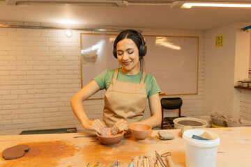 Happy Latin American woman creating pottery bowls wearing headphones