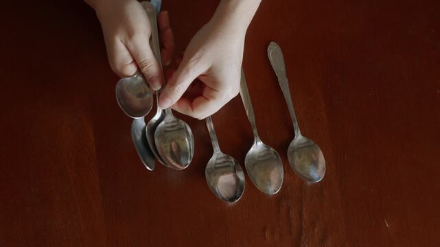 Top view of woman hands arranging several metal spoons on brown wooden table in home interior, concept of daily chores and organization.