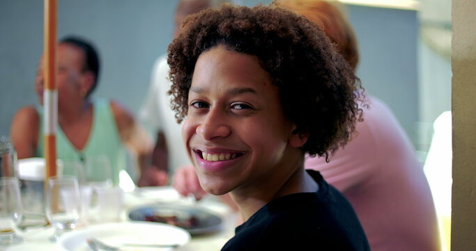 One Smiling young hispanic boy of African descent turns head towards camera, enjoying time at family gathering, seated at dining table surrounded by relatives, sharing joyful moments outdoors - Powered by Adobe