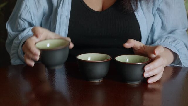 Female hands stacking three black ceramic cups with green interior on polished wooden table, natural daylight, symbolizing mindfulness, home order, simple living.