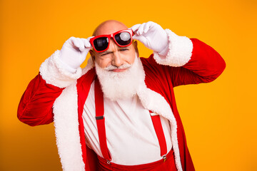 Santa in red festive suit adjusts bright red glasses against a warm orange background smiling holiday season beard hat suspenders