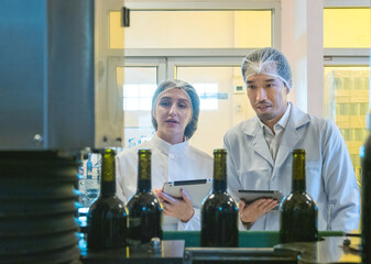 Two inspector wearing lab coat and hairnet check production detail on tablet in modern food...