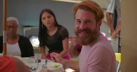 Bearded man smiling warmly at the camera while dining with friends in a backyard BBQ setting, with a young woman and older woman in the background, enjoying the gathering