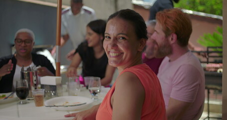 Woman smiling brightly at the camera during a lively family BBQ lunch, surrounded by friends and family enjoying an outdoor meal in a warm and cheerful atmosphere