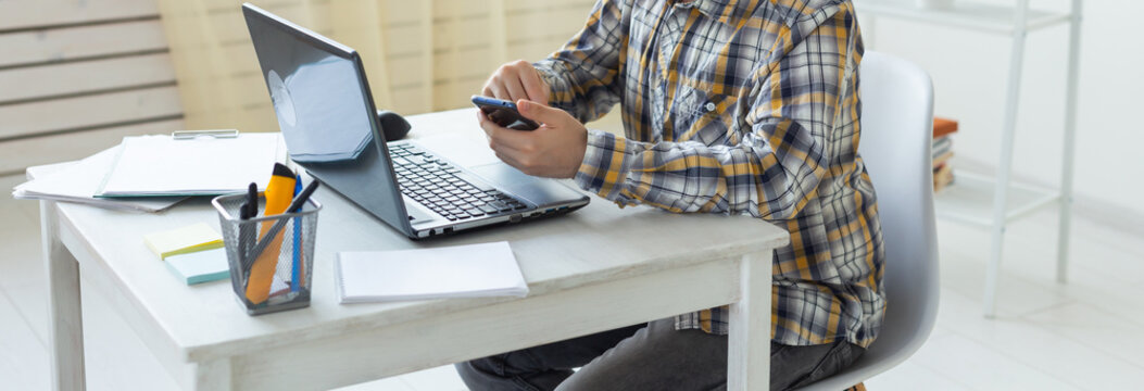 Male freelancer using smartphone while working on laptop at white desk. Banner with copy space. Multitasking, communication, and modern digital work concept.
