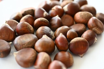 A pile of raw chestnuts displayed on a white surface in soft natural light. The smooth, glossy shells and warm brown hues emphasize the freshness and organic texture of the seasonal nuts.