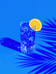 Tropical cocktail glass with lemon slice and ice cubes on bright blue background. Bold minimalist summer drink composition with palm shadow and vibrant contrast
