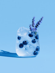 Refreshing blueberry and lavender mocktail with ice cubes in a transparent glass on bright blue background. Minimalist summer drink concept with vivid light and clean composition