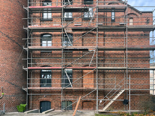 Historic red brick building under renovation with metal scaffolding covering the facade, multiple arched windows, and a paved area in front.