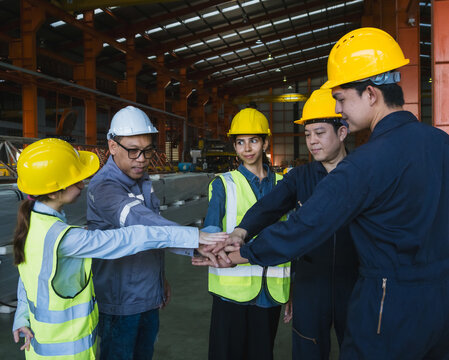 group five industrial worker wearing safety helmet and reflective vest standing circle inside factory placing their hands together center symbolize teamwork collaboration in safe working environment.
