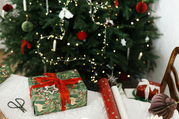 Merry Christmas and Happy Holidays! Wrapped christmas presents, modern ornaments, wrapping paper, ribbons on rustic table against decorated christmas tree with lights. Packing xmas gifts