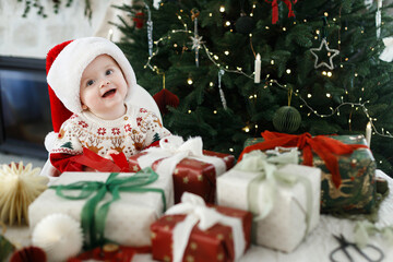 Merry Christmas! Happy cute baby in santa hat and festive sweater playing with christmas red bow on table with gifts against decorated christmas tree.