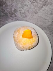 Close-up of a yellow/orange mango-flavored mochi ball, dusted with powder, topped with a piece of fresh mango. Focus highlights the soft, chewy texture of the sweet Japanese dessert.