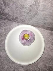 Close-up of a purple/grape-flavored mochi ball, powdered and topped with a slice of fresh red grape and white cream, served on a white plate. Soft, sweet Japanese dessert texture.