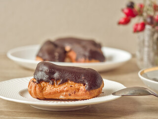 A single chocolate eclair on a white plate with a spoon, next to a white cup of coffee