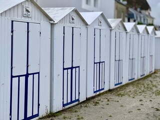 Fototapeta premium Row of traditional white and blue beach cabins on the sandy coast of Le Crotoy, Somme Bay, France, under a cloudy sky with tire tracks on the beach creating depth and leading perspective