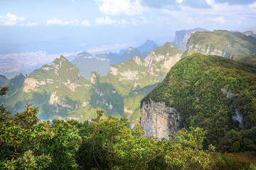 Aerial view of mountain cliffs covered with green forest and vegetation, showing rugged terrain with distant cityscape under a bright cloudy sky