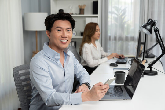 Profile smart Asian businessman looking camera to pose using smartphone at modern office. Blurry background woman colleague working on pc to check email marketing report or reply customer. Infobahn.
