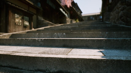 Steps worn smooth by time ascend toward a tranquil hillside location, surrounded by traditional buildings and cherry blossoms swaying softly in the breeze.