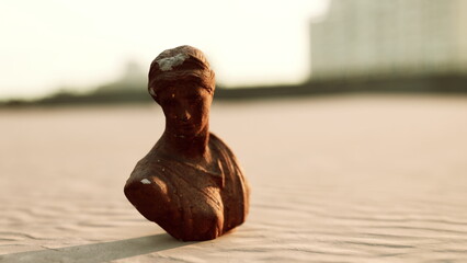 A weathered clay bust stands on a deserted beach, surrounded by soft sand. The warm glow of the sunset highlights the contours of the sculpture, creating a serene atmosphere. © icetray