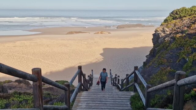 People walk down seaside stairs at dusk, Individuals descend wooden staircase towards sandy shores amid coastal cliffs and evening Sines Portugal