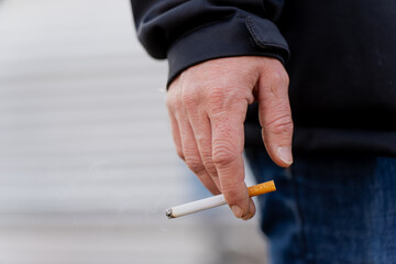 Adult male hand holding lit cigarette with visible smoke and ash tip showing personal habit, nicotine addiction theme and lifestyle concept in outdoor environment