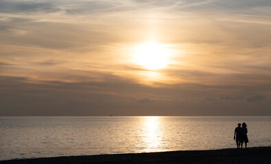 Fototapeta premium A man and a woman are standing on the shore of the sea, watching the sunset together. Both have their cell phones in their hands. The sun is setting behind the clouds, spreading red light.