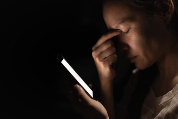 A tired woman holds her phone in a dark room, rubbing her eyes from stress and screen fatigue....