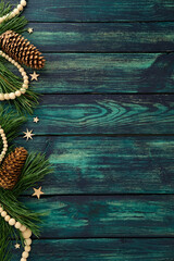 Rustic Christmas border with pine branches, pinecones, wooden beads, and stars on dark teal wooden background, festive frame with copy space.