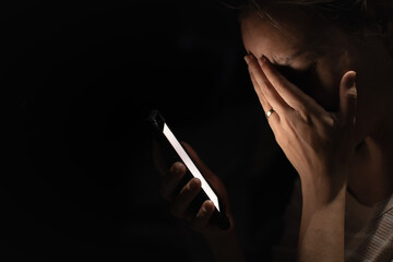 Woman crying while looking at her smartphone in the dark, covering her face in emotional pain....