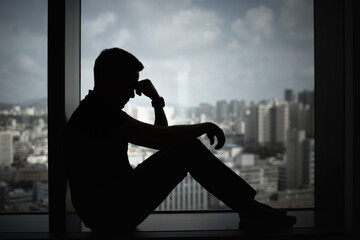 Silhouette of a man sitting by a window, holding his head in stress and sadness. Represents...