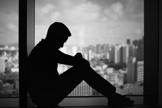 Silhouette of a man sitting by a window overlooking a city, showing sadness and emotional struggle. Represents depression, anxiety, isolation, and mental health issues