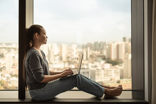A young woman works on her laptop while sitting by a large window overlooking the city. Represents remote work, freelance lifestyle, online business, and productivity

