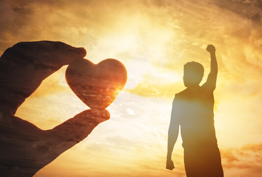 Inspirational image of a hand holding a heart shape and a silhouette celebrating at sunset. Symbolizes love, hope, strength, success, and positive energy