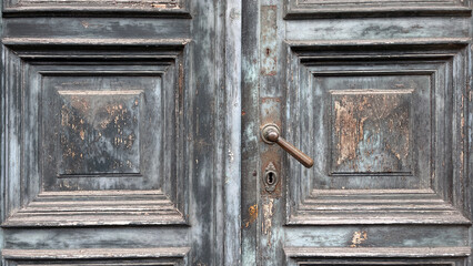 Close-up of a weathered wooden double door with ornate panels, peeling paint in shades of gray and blue, and a vintage metal handle with a keyhole, showing signs of age and patina.