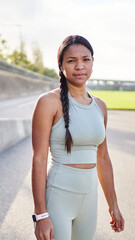 Vertical. Calm African millennial woman standing outdoors in park after exercise, wearing sportswear and looking confident at camera. Natural light portrait shows strength balance and active lifestyle